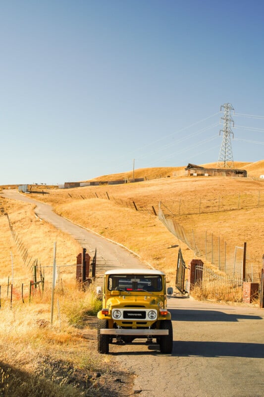 A yellow vintage off-road vehicle is parked on a winding road surrounded by dry, golden grass fields. In the background, there's a power line tower and a wooden structure under a clear blue sky.