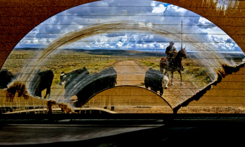 View through a dusty windshield of a cowboy on horseback herding cattle across a rural dirt road. The windshield has a clear arc surrounded by dirt. The sky is filled with scattered clouds above the expansive landscape.