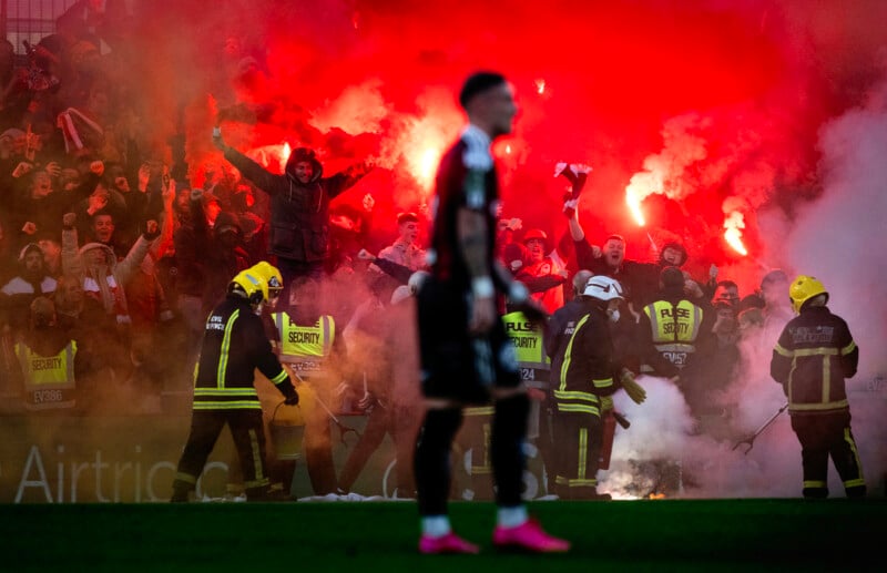A crowd of football fans cheer behind a barrier, holding flares that create a red smoky haze. Security personnel and firefighters stand nearby. The silhouette of a player is visible in the foreground.