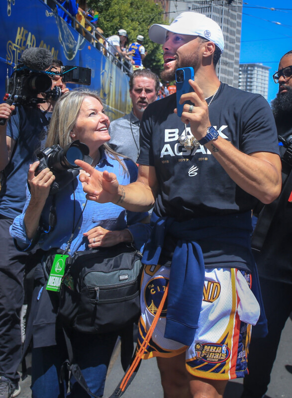 A joyful man in a black "Back Again" shirt interacts with a smiling woman holding a camera. They're surrounded by media personnel on a bustling urban street. He's wearing colorful shorts and a cap, and there are spectators and buildings in the background.