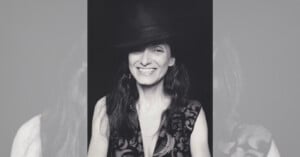 A woman with long hair smiles, tilting her head slightly. She wears a dark, wide-brimmed hat and a patterned sleeveless top with a deep neckline. The photo is in black and white, with the woman centered against a plain background.