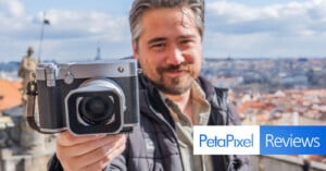 A man holds a large, vintage-style camera up to the viewer. He stands outdoors with a cityscape in the background. The bottom of the image displays "PetaPixel Reviews" logos.