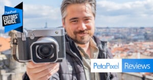 A man outdoors holds a silver camera towards the viewer, smiling. The background shows a scenic cityscape. "Petapixel Editors' Choice" and "Petapixel Reviews" graphics appear on the image.