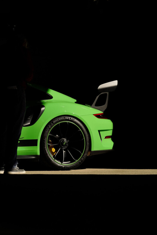 A vibrant green sports car is partially visible, illuminated against a dark background. The focus is on the sleek rear wheel and spoiler, with a person standing nearby. The tire has branding, and the scene is dramatically lit.
