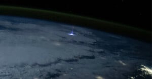 A stunning view from space showing the Earth's horizon at night, with a luminous blue burst of light piercing the cloud cover, likely a lightning event. The distant city lights are visible in contrast with the dark, expansive sky.