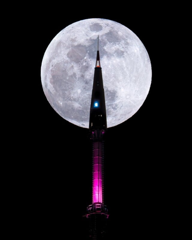 A tall, illuminated tower with a sharp spire stands against the night sky. The large, bright full moon appears directly behind the spire, creating a dramatic silhouette effect. The tower is lit with a magenta hue.