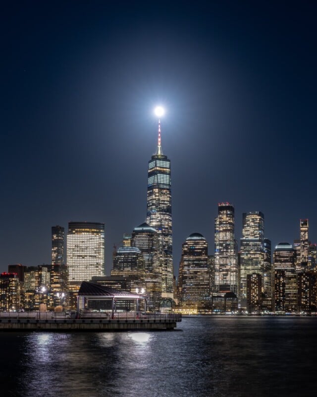 A city skyline at night, illuminated by bright lights. A tall skyscraper, topped with a glowing light, stands prominently among other buildings. The sky is dark, and the scene reflects off a body of water in the foreground.