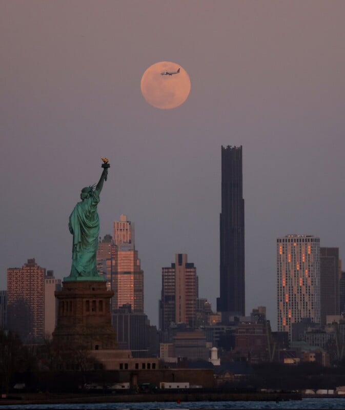 The Statue of Liberty is in the foreground with the New York City skyline behind it. A large full moon is visible in the sky, with an airplane silhouetted against it. The sky is dusky with a soft gradient of colors.