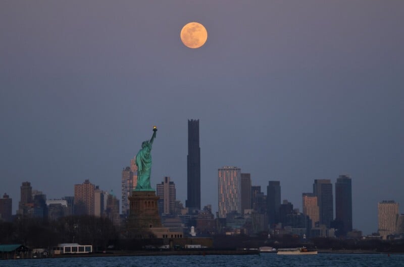 The Statue of Liberty is silhouetted against a city skyline at dusk, with a large full moon rising above skyscrapers in the background. The scene is set over a body of water, capturing a serene and majestic urban landscape.