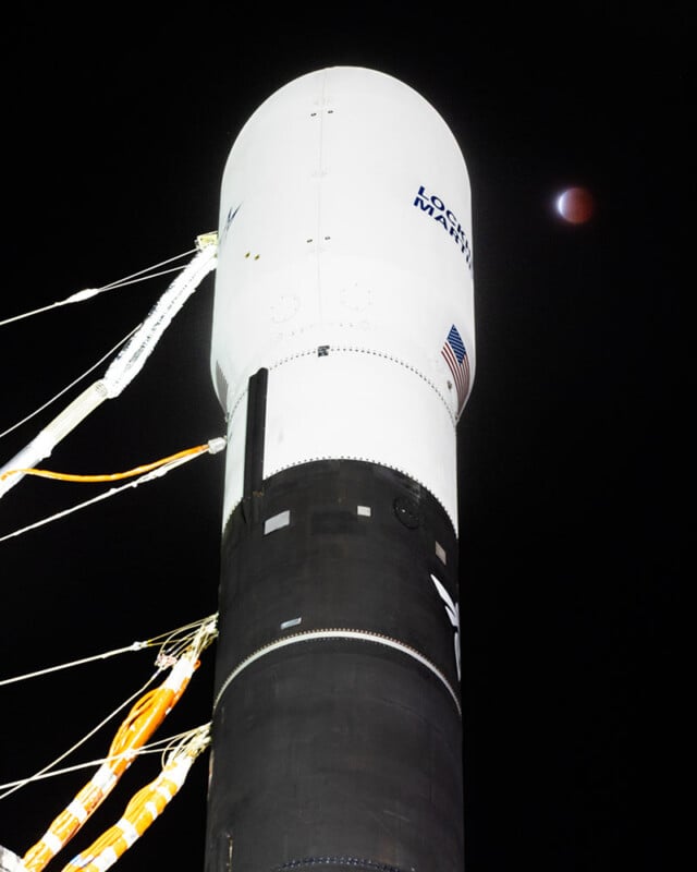 A white Lockheed Martin rocket stands tall against a dark night sky. Visible on the rocket are an American flag and company logo. In the background, there's a red-tinted moon partially obscured by shadow.