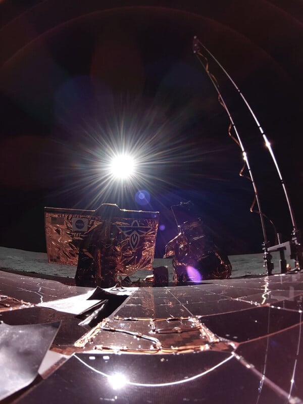 The image shows a spacecraft on a lunar surface with the sun shining brightly in the background. The spacecraft components are covered in reflective material, and the lunar horizon is visible under a dark sky.