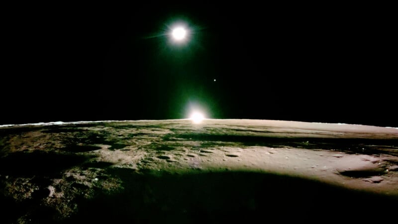 A stark, cratered lunar surface under a dark sky. Two bright celestial bodies, likely the Sun and Earth, appear above the horizon, casting light across the barren landscape.