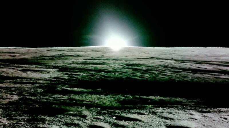 The image shows a bright light emerging over the horizon of a barren, rocky landscape, resembling the surface of a celestial body like the moon, against a dark sky. The foreground is textured with shadows.