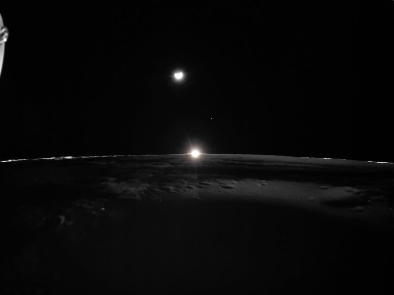 A dimly lit lunar surface with a bright glare on the horizon, likely from the sun peeking over. The Earth is visible in the dark sky above, alongside a smaller celestial body. The scene is mostly in shadow.