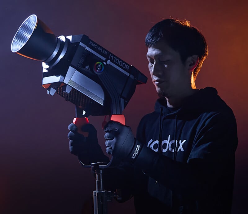 A person in a dark hoodie holds a large, professional camera light with "KNOWLED" and "M1000R" labels. The background is a gradient of orange and blue, creating a dramatic lighting effect. The individual is focused on adjusting the equipment.