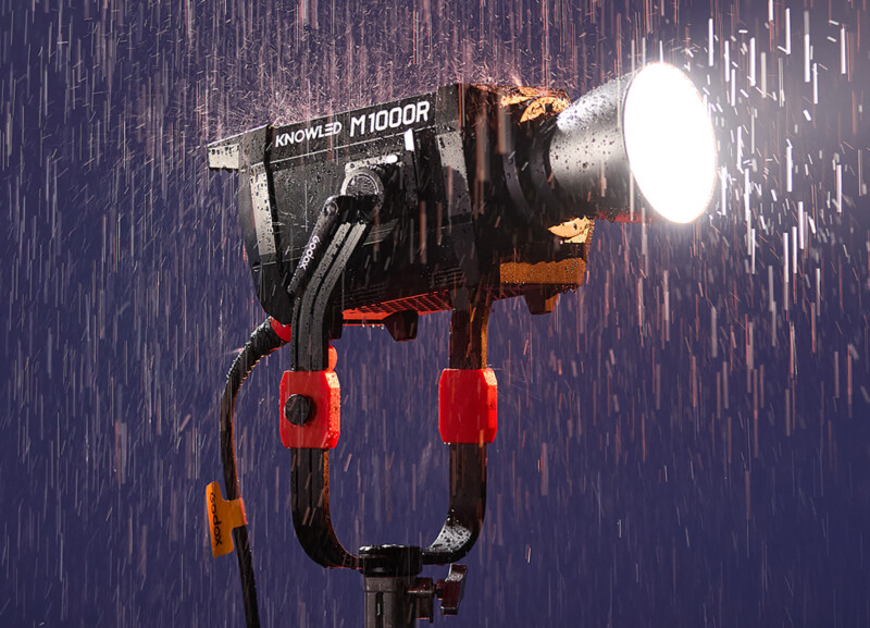 A powerful light stands on a tripod, illuminated and covered in raindrops during heavy rain. The light is labeled "Knowled M1000R" and has red and black components, contrasting against a dark blue background.