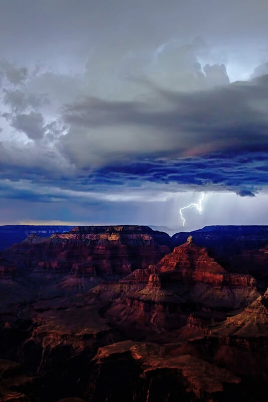 A dramatic view of the Grand Canyon at dusk with dark storm clouds overhead. A bright flash of lightning illuminates the sky, casting shadows over the rugged canyon landscape.