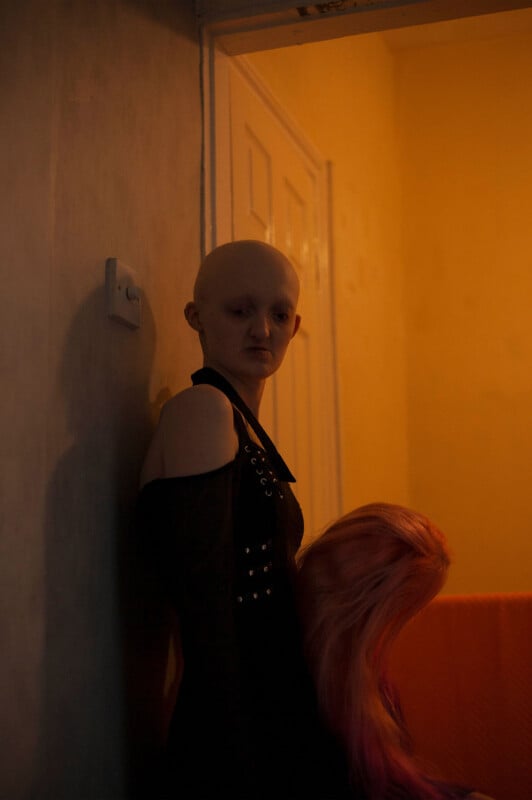 A person wearing a dark off-shoulder outfit stands against a wall in a dimly lit room. They have no hair and are looking down. Nearby, a bright orange object, possibly a wig, rests on a surface. Warm, yellowish light fills the space.