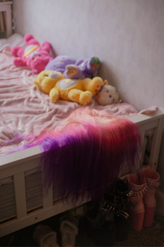 Kids' bed with pink and purple plush toys and a blanket. A colorful, long wig or hairpiece drapes over the edge. Pink boots with black studs are on the floor next to the bed, showing a whimsical and playful room setting.