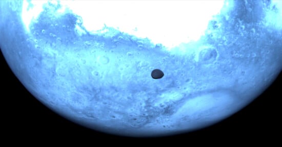 A close-up view of a blue-tinted planet surface with craters and rough terrain. A small, dark circular shadow is visible on the planet's surface, indicating an eclipse or transit event.