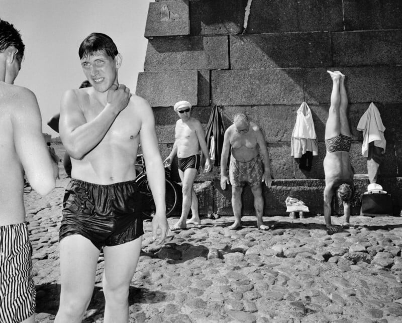 A group of shirtless men in swim trunks stand on a cobblestone area by a large stone wall. One man walks towards the camera, another balances on his hands, and others are standing or adjusting clothes hanging on the wall.