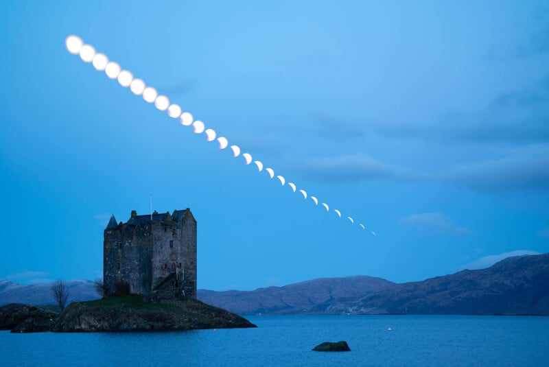 A serene evening scene with an old stone castle on a small island, surrounded by calm blue waters. A sequence of moon phases is captured in the sky, forming an arc as they appear to set behind distant hills.