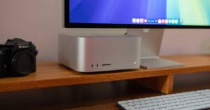 A desktop setup featuring a silver computer, a monitor displaying a colorful screen, a camera on the left, and a white keyboard on a wooden desk.