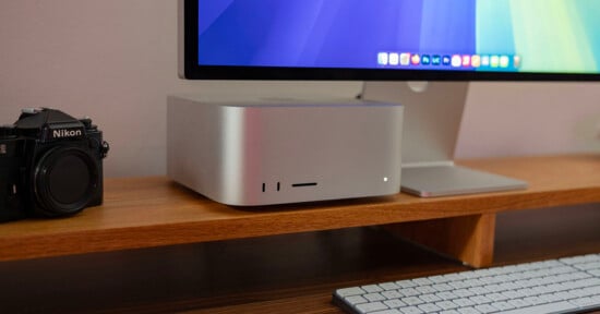 A desktop setup featuring a silver computer, a monitor displaying a colorful screen, a camera on the left, and a white keyboard on a wooden desk.