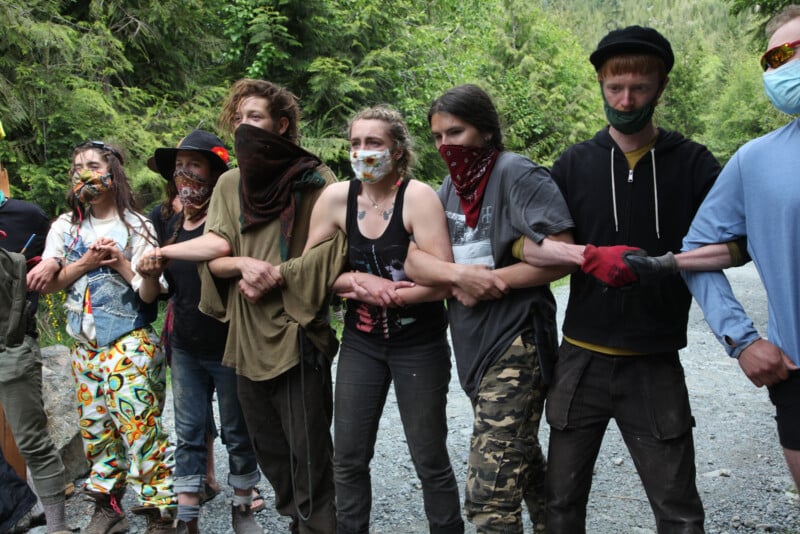 A group of people stands in a line, arm in arm, blocking a gravel path. They wear casual clothing, some with masks and bandanas covering their faces. The background is a lush forest.