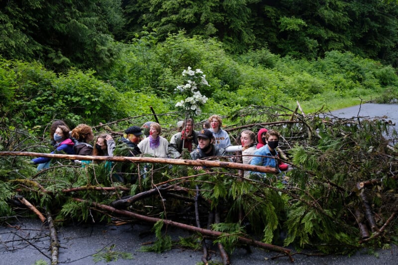 A group of people stand behind a barricade made of branches and foliage on a road surrounded by dense greenery. They appear to be participating in a protest, with some wearing colorful clothes and masks.