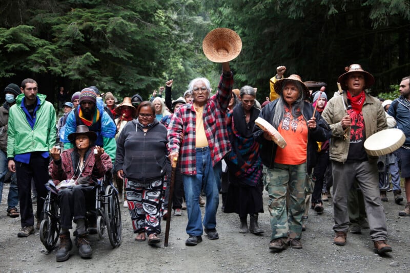 A group of people walks along a forest path. An elderly man raises a woven hat, and a woman next to him holds a drum. Some wear traditional attire, including blankets and beadwork. The background shows tall trees, suggesting a forest setting.