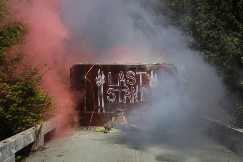 A large barricade with the words "LAST STAND" in white, set on a forested path. Red and gray smoke surround it, and greenery is visible on both sides. A person sits at the base of the barricade, partially obscured by the smoke.