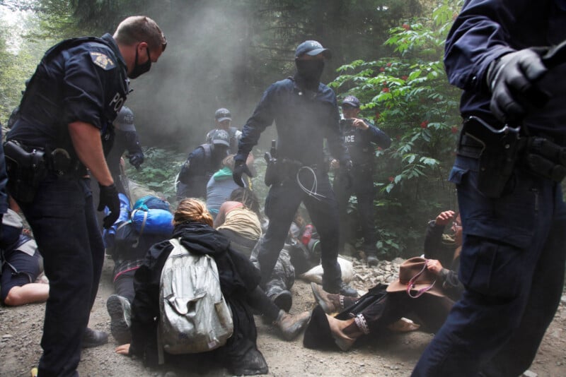 Police officers in protective gear detain a group of protesters on a dirt path in a wooded area. Some officers stand over the protesters, who are seated and lying on the ground, with smoke or dust in the air.