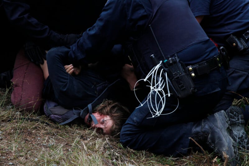 A person is lying on the ground with hands restrained, surrounded by several police officers. One officer is pressing the person down. The scene is outdoors on grass, and the person appears distressed. Officers have equipment on their belts.