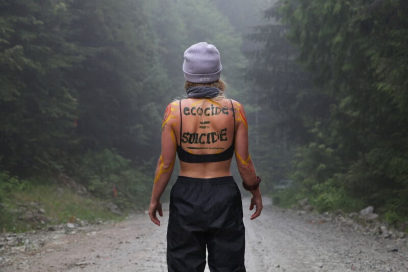 A person with long hair stands on a misty forest path, facing away. They wear a beanie, sports bra, and pants. Words "ECOCIDE = SUICIDE" are painted on their back. The scene is surrounded by dense green trees and a gravel path.