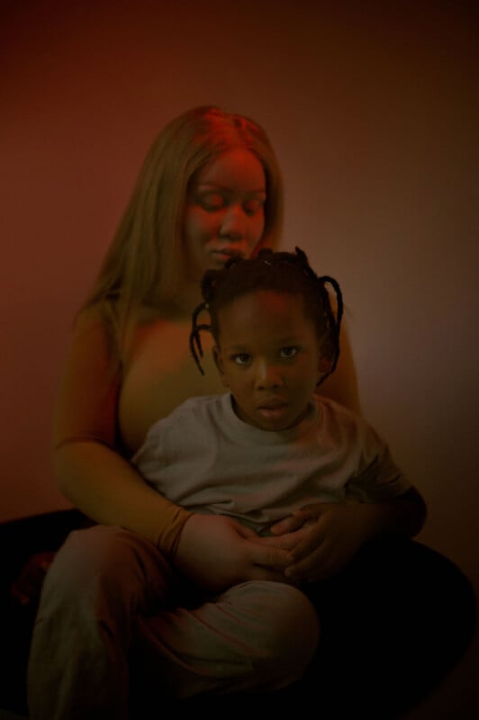 A woman with long hair sits holding a child on her lap. The child, with short braided hair, looks towards the camera. The lighting is warm and dim, creating a soft atmosphere against a plain background.