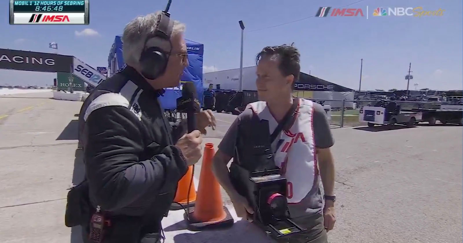 A TV presenter in a racing suit holds a microphone, speaking to a man with a camera around his neck. They are outdoors at a racetrack with tents and cars in the background. Various logos and time information are displayed on the image.