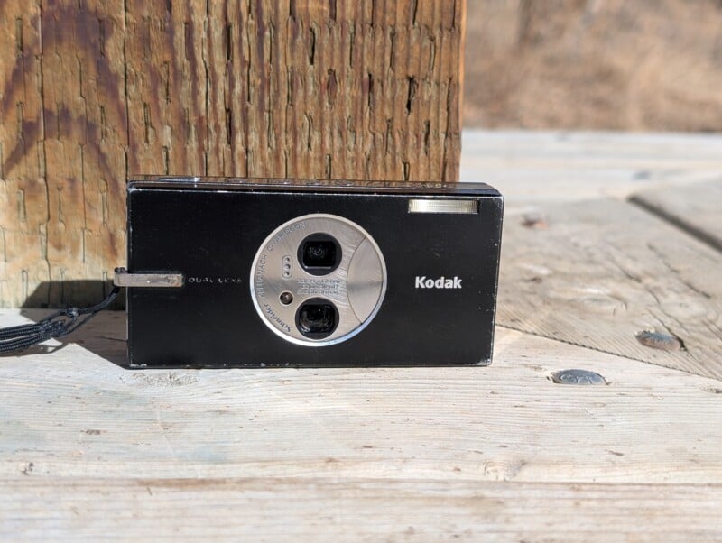 A black Kodak compact camera with dual lenses sits on a wooden surface, leaning against a wooden post. The sunlight casts a shadow on the camera, highlighting its glossy finish and silver circular lens area.