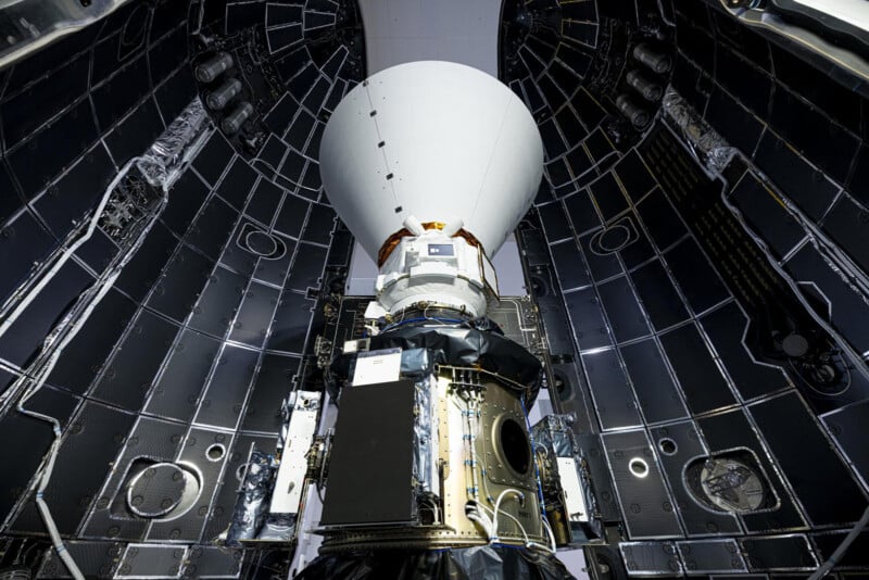 A spacecraft is positioned inside a rocket fairing. The interior walls of the fairing are lined with dark panels. The spacecraft has a white, conical top and various electronic equipment visible on its body.