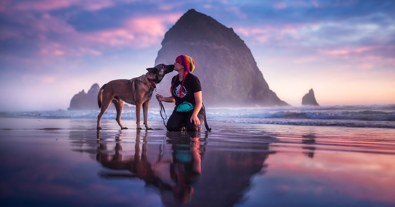 A person kneels on a reflective beach as a dog affectionately licks their face. The sky is filled with pink and blue hues at sunset, and a large rock formation rises in the background amidst gentle ocean waves.