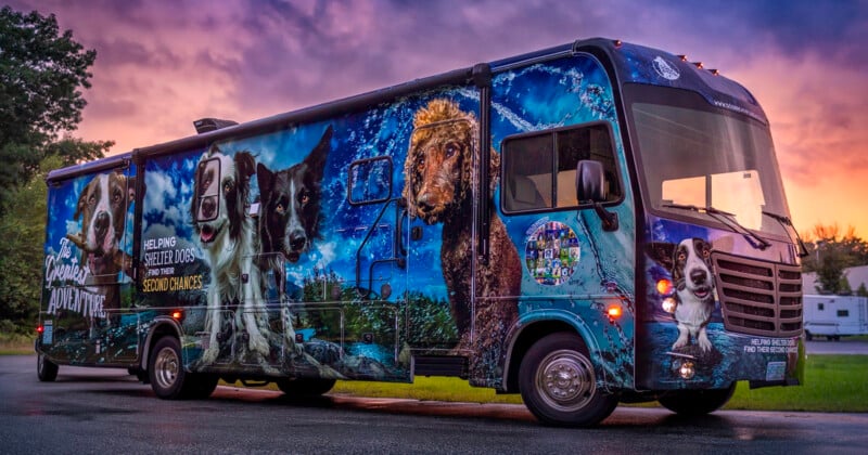 A large RV with a colorful mural featuring dogs and nature scenes parked on a road. The sky is a vibrant mix of purple, orange, and pink hues at sunset. The side of the RV displays text supporting dog rescue and second chances.