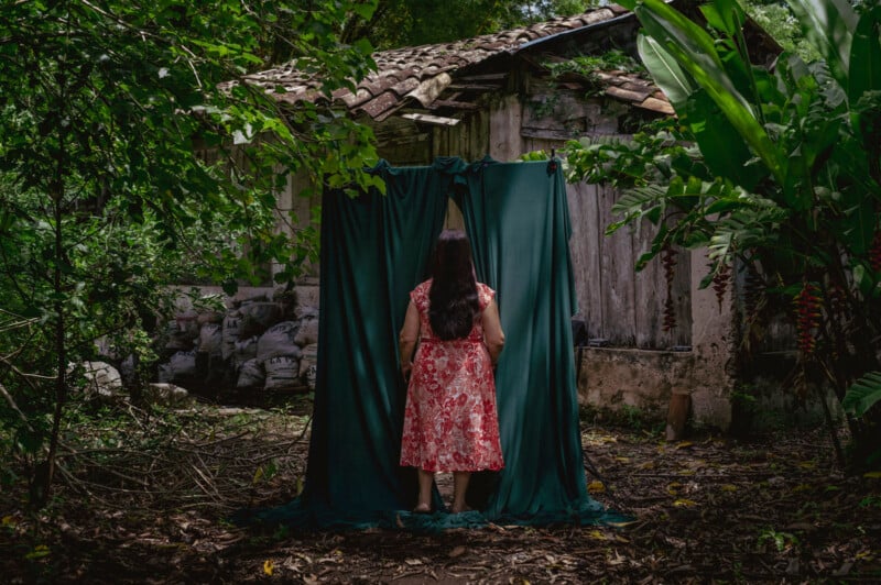 A woman in a red floral dress stands facing away, looking at a small, old building. She is behind a dark green curtain hanging outdoors, surrounded by lush greenery and large leaves. The scene conveys a mysterious, secluded atmosphere.