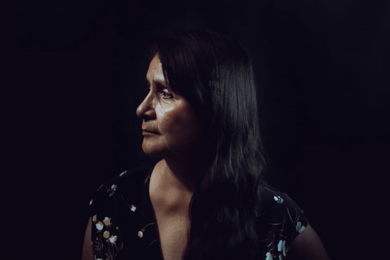 A woman with long dark hair is shown in profile against a dark background. She is wearing a floral-patterned blouse, and the lighting highlights her face, giving a dramatic effect.