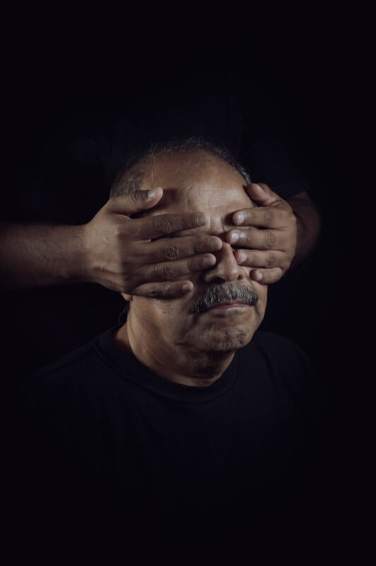 A man with a mustache sits with closed eyes as another person's hands cover them from behind. The background is dark, creating a dramatic contrast that highlights the faces and hands.