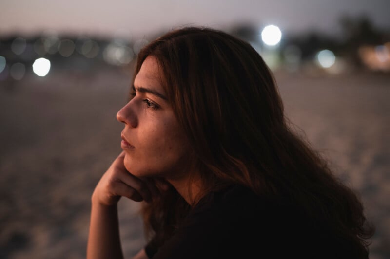 A person with long brown hair gazes thoughtfully into the distance while sitting on a sandy beach during twilight. Blurred lights twinkle in the background, creating a serene and contemplative atmosphere.