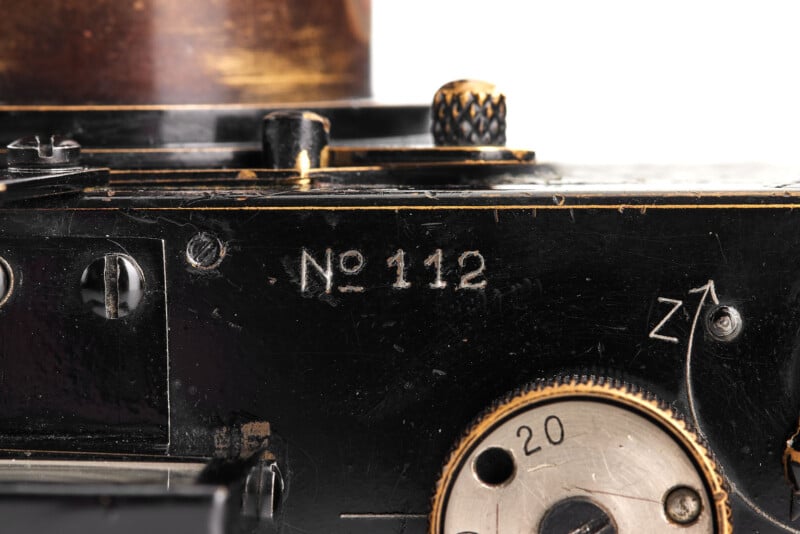 Close-up of a vintage black camera with visible brass details. The text "No 112" is engraved on the surface. A dial with the number 20 and a "Z" with an arrow can also be seen. The metal has a worn appearance.