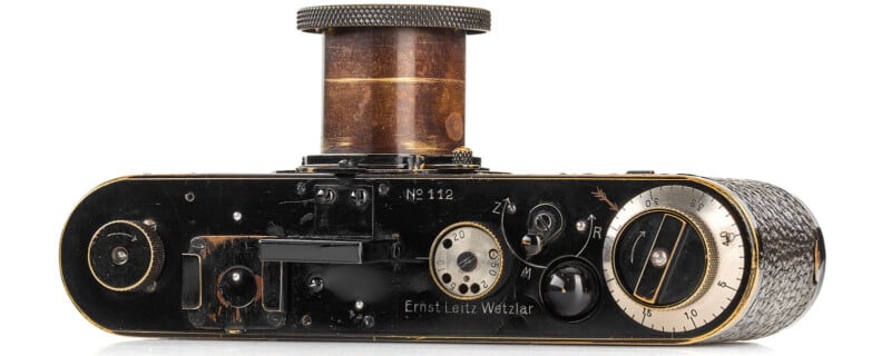 A vintage camera with the lens extended. The bottom view shows dials and markings, including "Ernst Leitz Wetzlar" and "No. 112." The design features a textured grip and brass-like finish, indicating an early 20th-century model.