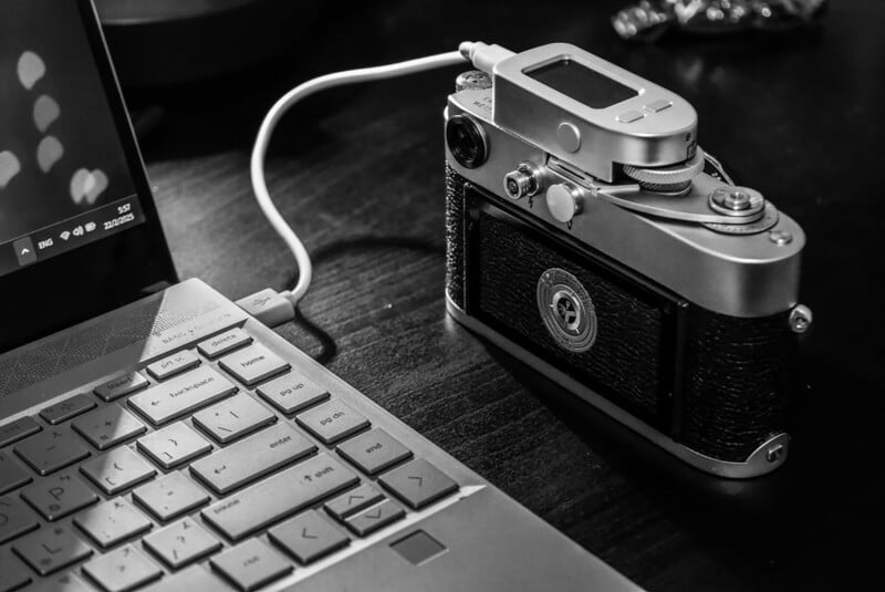 Black and white image of a vintage camera connected to a laptop via a cable. The camera sits on a dark surface, and part of the laptop keyboard is visible.