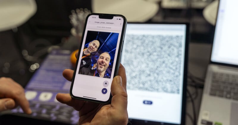 A person holds a smartphone displaying a selfie of two individuals smiling. In the background, there's a laptop with a QR code visible on the screen. Other devices and a person's hand are also partially visible.