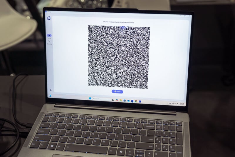 A laptop screen displaying a large QR code in the center. The laptop is on a table, and the keyboard and trackpad are visible below the screen. The screen has a white background with some blue elements.